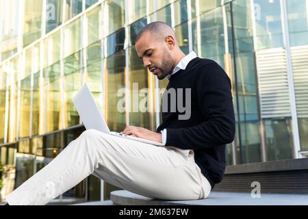 Side view of focused black man in smart casual clothes typing information on netbook while working on business project outdoors Stock Photo