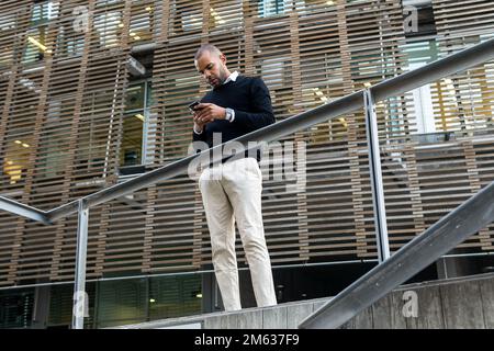 Low angle of serious ethnic man in smart casual wear browsing mobile phone surfing internet during break from work standing outside building Stock Photo