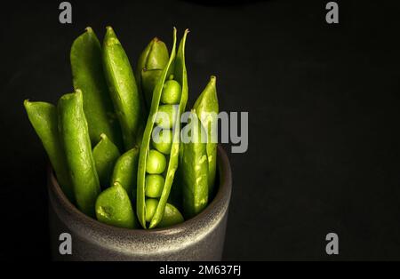 Bunch of ripe green pods with fresh peas placed in ceramic pot against black background Stock Photo