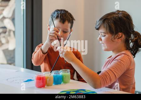Happy girl and boy washing paintbrushes in cups with water while sitting at table and painting environmental posters in daytime at home Stock Photo