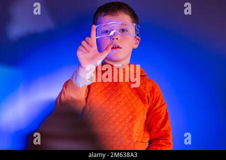 Kid in orange smart goggles experiencing digital cyber world while standing in dark studio with blue light Stock Photo