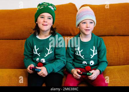 Brother and sister in similar funny sweaters and warm hats sitting on sofa and playing videogame together on Christmas day at home Stock Photo