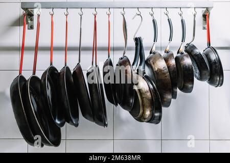 Various sized metal pans hanging in row on hooks attached to railing near white tiled wall Stock Photo