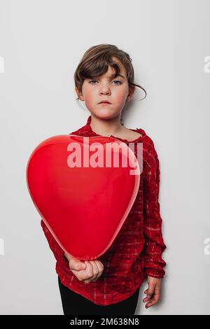 Serious girl showing heart shaped balloon of red color and looking at camera against gray wall on Saint Valentine Day Stock Photo