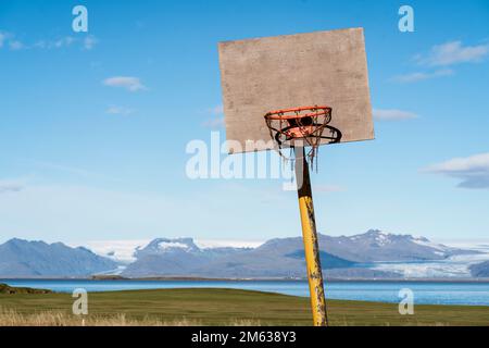 Basketball hoop placed on ground near calm blue lake and mountains under bright sky on sunny day in Iceland Stock Photo