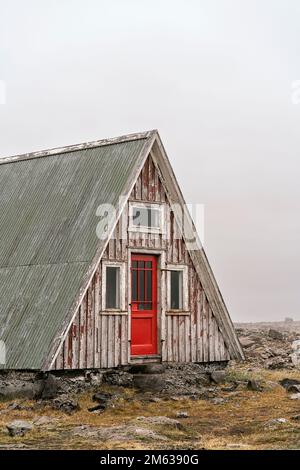 Exterior of old shabby wooden A frame house with metal roof and red door located on rocky terrain under gloomy sky Stock Photo