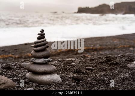 Pile of rocks stacked together on black volcanic sandy beach near wavy ocean against cloudy sky in Iceland Stock Photo
