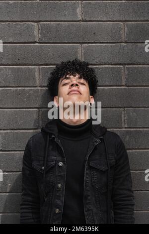 Young Latin American male with curly hair in black jacket leaning on brick wall with closed eyes Stock Photo