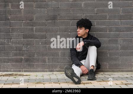 Full body of young ethnic male migrant in casual outfit sitting on paved ground having opportunities for working and studying after immigration from V Stock Photo