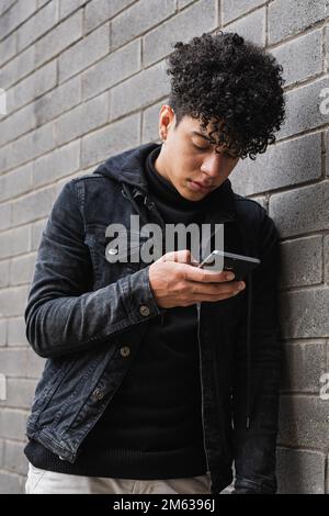 Young curly haired male in stylish outfit standing near brick wall messaging on mobile phone while living in Colombia after immigration from Venezuela Stock Photo