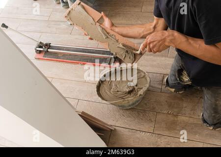 High angle of crop anonymous male master in black t shirt and dirty pants sitting on floor and applying cement on piece of tile with putty knife durin Stock Photo