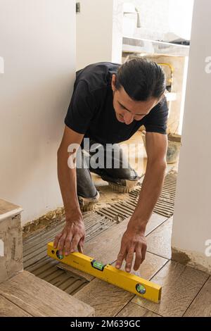 High angle of adult male worker in black t shirt sitting on floor and installing carpenter level on freshly laid tiles in apartment Stock Photo