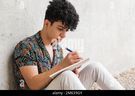 Positive focused Venezuelan male in casual clothes sitting on concrete ground while writing thoughts in notebook Stock Photo