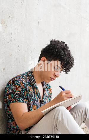 Positive focused Venezuelan male in casual clothes sitting on concrete ground while writing thoughts in notebook Stock Photo