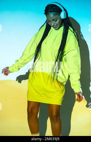 Young smiling African American woman with Afro braids in bright hoodie dress standing against blue and yellow lights while dancing and listening to mu Stock Photo