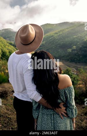 Back view of young female in romantic dress and man in casual outfit and straw hat embraced while enjoying mountains Stock Photo