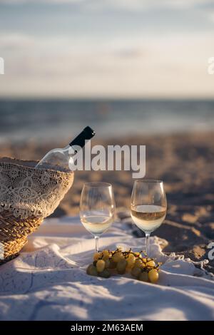 Bunch of white grapes and glasses of wine placed on blanket near basket with bottle on blurred background of sea and beach Stock Photo