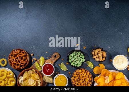 Various unhealthy salty bar snacks set. Dark grey table background with traditional party snacks - chips, onion rings, salted nuts, crisps,  pretzels Stock Photo