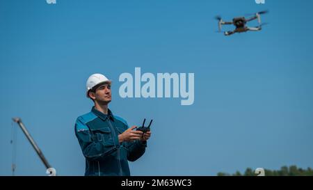 A man in a helmet and overalls controls a drone at a construction site. The builder carries out technical oversight. Stock Photo