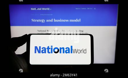 Person holding mobile phone with logo of British media company National World plc on screen in front of business web page. Focus on phone display. Stock Photo
