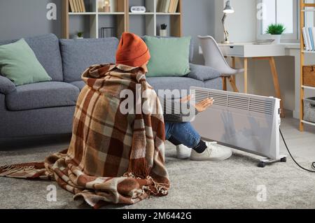 Young woman is trying to warm up by an electric heater in a cold house in winter Stock Photo