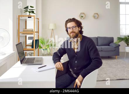 Successful man with beard and mustache looks at camera sits at desk with laptop in home office Stock Photo