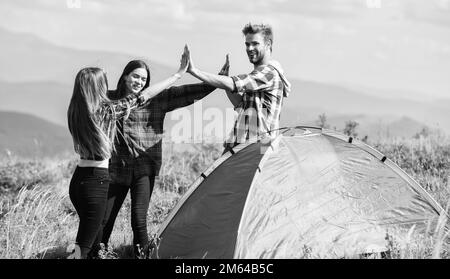 Good job. Teamwork concept. Hiking activity. Friends set up tent on top mountain. Camping equipment. Weekend in mountains. Man and girls having fun in Stock Photo