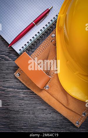 Leather tool belt hard hat checked notepad ball-point pen on wooden board construction concept. Stock Photo