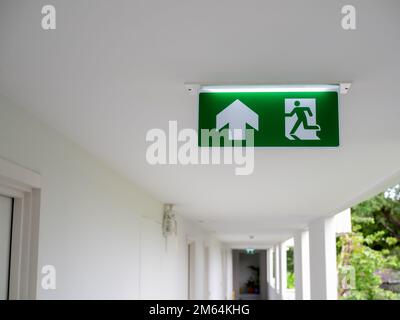 Green fire escape sign hang on the ceiling at the corridor in the white hotel building near the stairway. Emergency fire exit sign, warning plate with Stock Photo