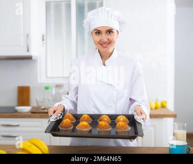 Cheerful female chef in white uniform holding baking tray of muffins Stock Photo