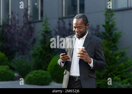 Mature businessman in business suit using phone outside office building, african american boss holding cup of hot drink, smiling typing text message and browsing websites. Stock Photo