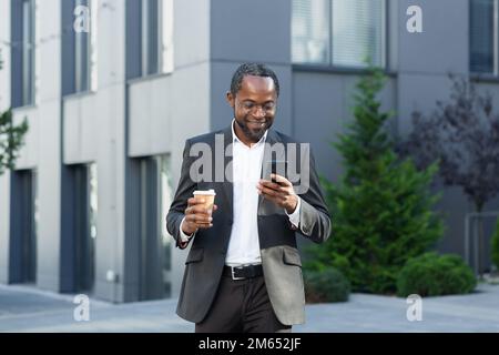 Mature businessman in business suit using phone outside office building, african american boss holding cup of hot drink, smiling typing text message and browsing websites. Stock Photo