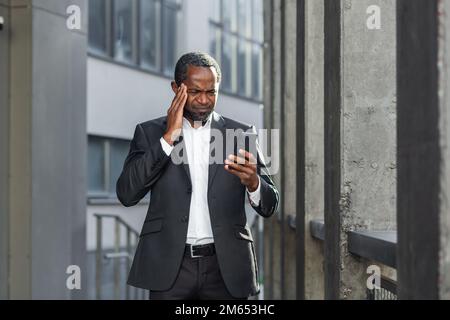 African american boss outside office building with phone in hands reading bad news online, man received notification about money loss and fraud, upset businessman. Stock Photo