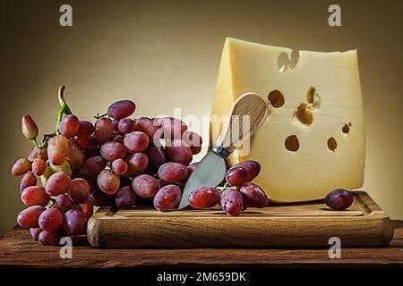 big piece of cheese with rose grape and little knife on wooden board yellow gradient background Stock Photo