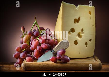 big piece of cheese with rose grape and little knife on wooden board brown gradient background Stock Photo