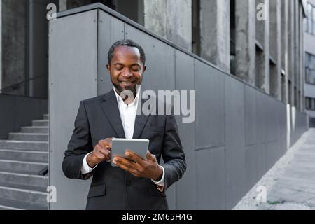 Successful african american man in business suit smiling and using tablet computer, businessman outside modern office building walking. Stock Photo