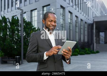 Successful african american man in business suit smiling and using tablet computer, businessman outside modern office building walking. Stock Photo