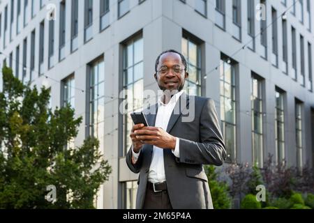 Portrait of African American mature businessman, senior man outside office building holding phone in hands smiling and looking at camera. Stock Photo