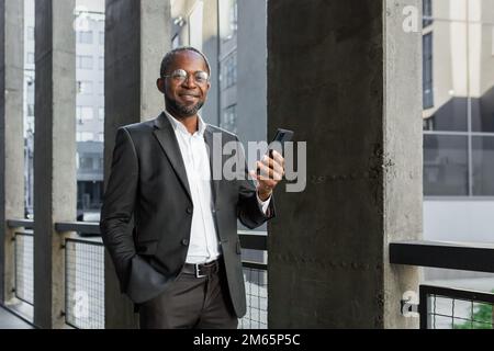 Portrait of African American mature businessman, senior man outside office building holding phone in hands smiling and looking at camera. Stock Photo