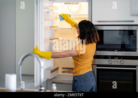 Young Housewife Wiping Shelves In Empty Fridge While Doing Cleaning In Kitchen Stock Photo