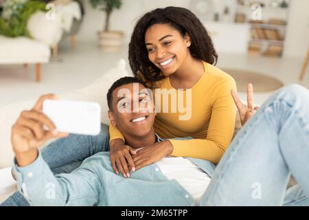 Smiling millennial african american wife hugging husband, relaxing on sofa and taking selfie Stock Photo