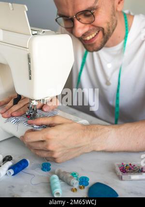A smiling blond man in glasses is sewing in home workshop. A seamster with a measuring tape on his neck in a white t-shirt. Concept of small business Stock Photo