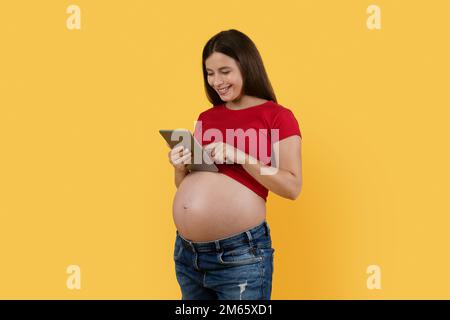 Smiling Pregnant Woman Using Digital Tablet While Standing Over Yellow Studio Background Stock Photo