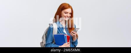 Lovely good-looking female with red hair, heading to college, holding backpack, notebooks smiling as answer friend message, chatting glancing mobile Stock Photo