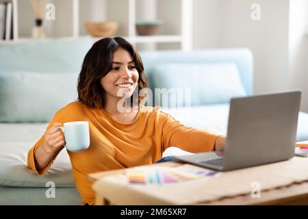 Distance Work. Smiling Arab Female Freelancer Drinking Coffee And Working With Laptop Stock Photo