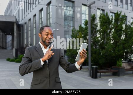 Mature african american boss businessman outside office building with headset and tablet talking cheerfully on video call, man in business suit remote chat with colleagues. Stock Photo