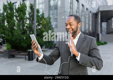 Mature african american boss businessman outside office building with headset and tablet talking cheerfully on video call, man in business suit remote chat with colleagues. Stock Photo
