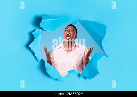 Angry oversize black woman shouting freaking out posing in hole of torn paper on blue studio background Stock Photo