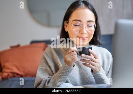 Close up portrait of beautiful asian woman drinks her coffee, smells drink in cup, takes brake from working or studying on laptop Stock Photo
