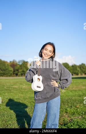 Cheerful female guitar player has pink hair poses with musical instrument  wears orange jacket entert Stock Photo by wayhomestudioo
